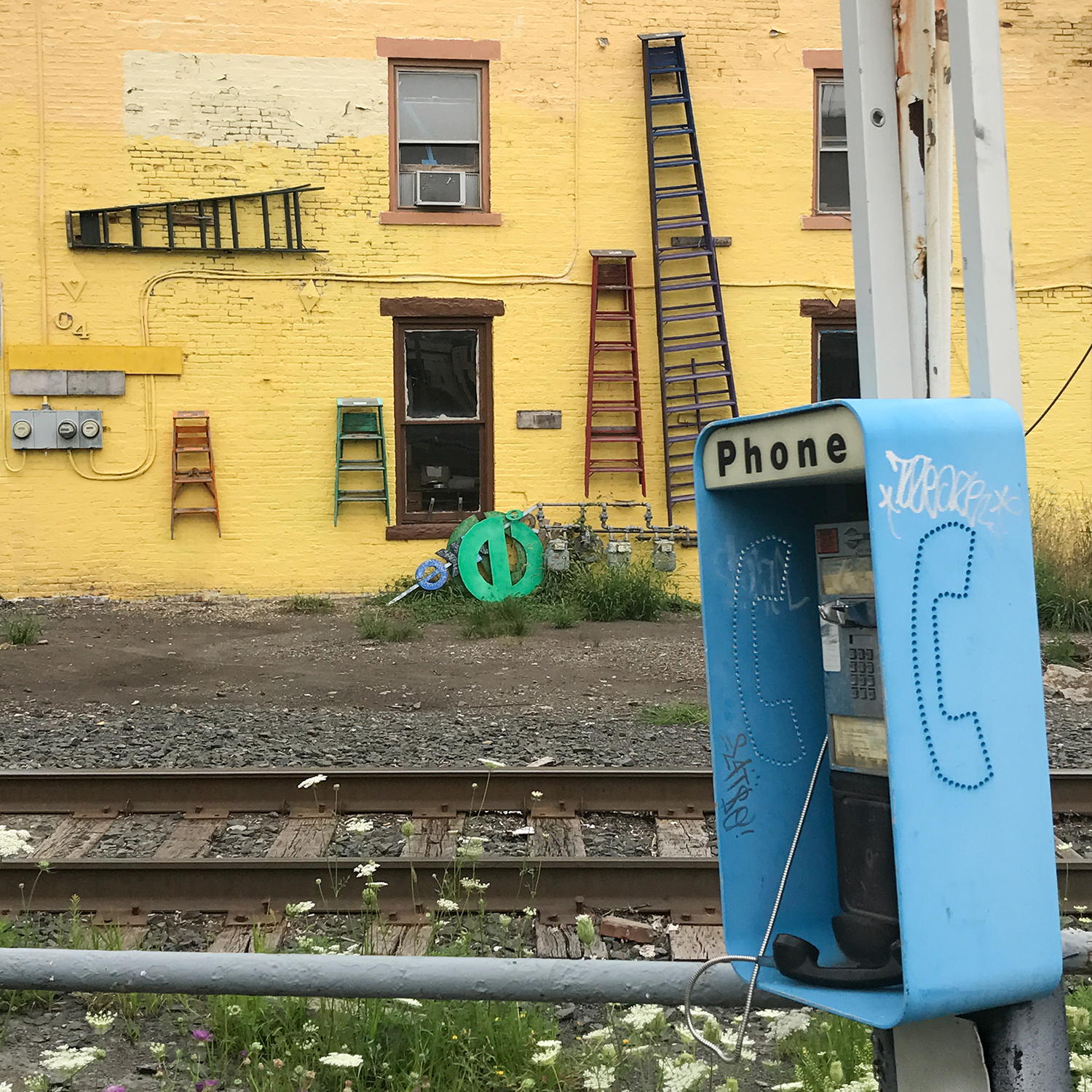 Amy Becker Ladders (detail), 2017 Archival pigment print 10 x 7 ½ inches. It depicts a blue payphone with some graffiti. Behind it sits train tracks and a yellow wall covered in windows and ladders.