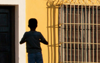 A young child standing in between a caged window and a door. Nancy Ori An Afternoon Memory, 2017 Archival inkjet photograph 18 x 18 inches