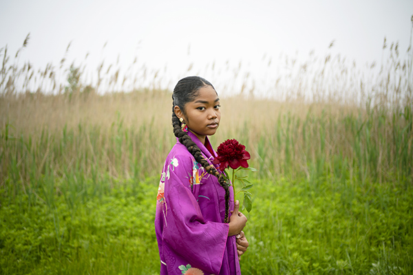 Hidemi Takagi, Mikua from the "IDENTITIES" series, 2022, Digital C Print on the textile. Shows a colored girl wearing purple. She has large, colorful earrings and is holding a red flower.