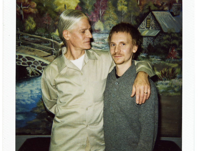 Davi Russo, Frame 60. 2007 from the series Picture Time, 1987-2007, Polaroid, 3.5 x 4.2 inches, courtesy of the artist. It displays two men. The one on the left puts his arm around and looks at the one on the right, who is focused on the camera. Behind them is a mural of a forest scene, which also has river with a bridge and a cottage.