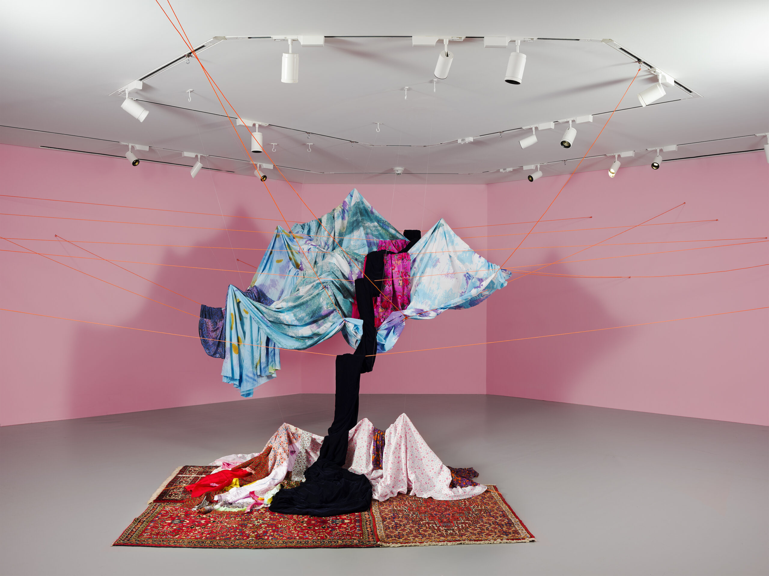 A contemporary art installation in a gallery with pink walls, featuring a central structure made of draped and suspended colorful fabrics in shades of blue, pink, black, and floral prints. The fabric is anchored to the ceiling and walls with taut orange cords, and some pieces cascade onto patterned rugs on the floor below.