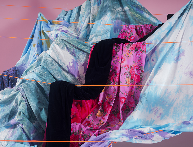 A close-up view of the fabric installation, showing layered textiles in vibrant hues of blue, purple, pink, and black, including floral and abstract patterns. The fabrics are suspended and partially gathered, intersected by thin orange cords creating tension across the composition.