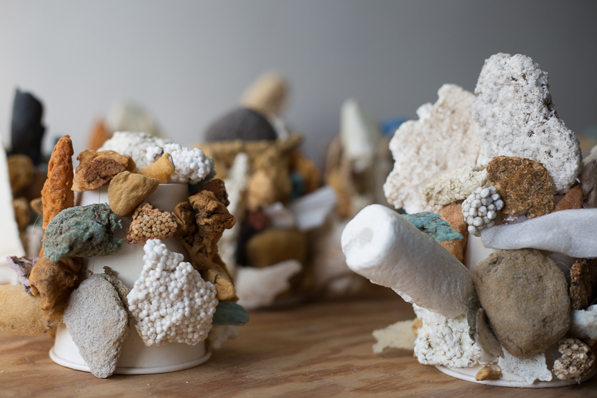 Close-up of an abstract sculptural arrangement made from assorted natural and artificial materials attached to white paper cups. The objects include porous rocks, foam, sponge-like fragments, Styrofoam, and earthy-toned stones in shades of brown, white, gray, and muted turquoise. The textures range from rough and jagged to smooth and rounded, creating a clustered, coral-like appearance on a wooden surface with other similar sculptures blurred in the background.