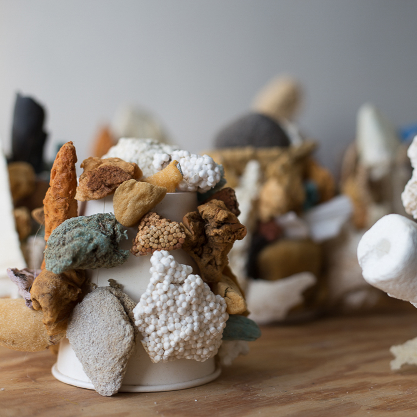Close-up of an abstract sculptural arrangement made from assorted natural and artificial materials attached to white paper cups. The objects include porous rocks, foam, sponge-like fragments, Styrofoam, and earthy-toned stones in shades of brown, white, gray, and muted turquoise. The textures range from rough and jagged to smooth and rounded, creating a clustered, coral-like appearance on a wooden surface with other similar sculptures blurred in the background.