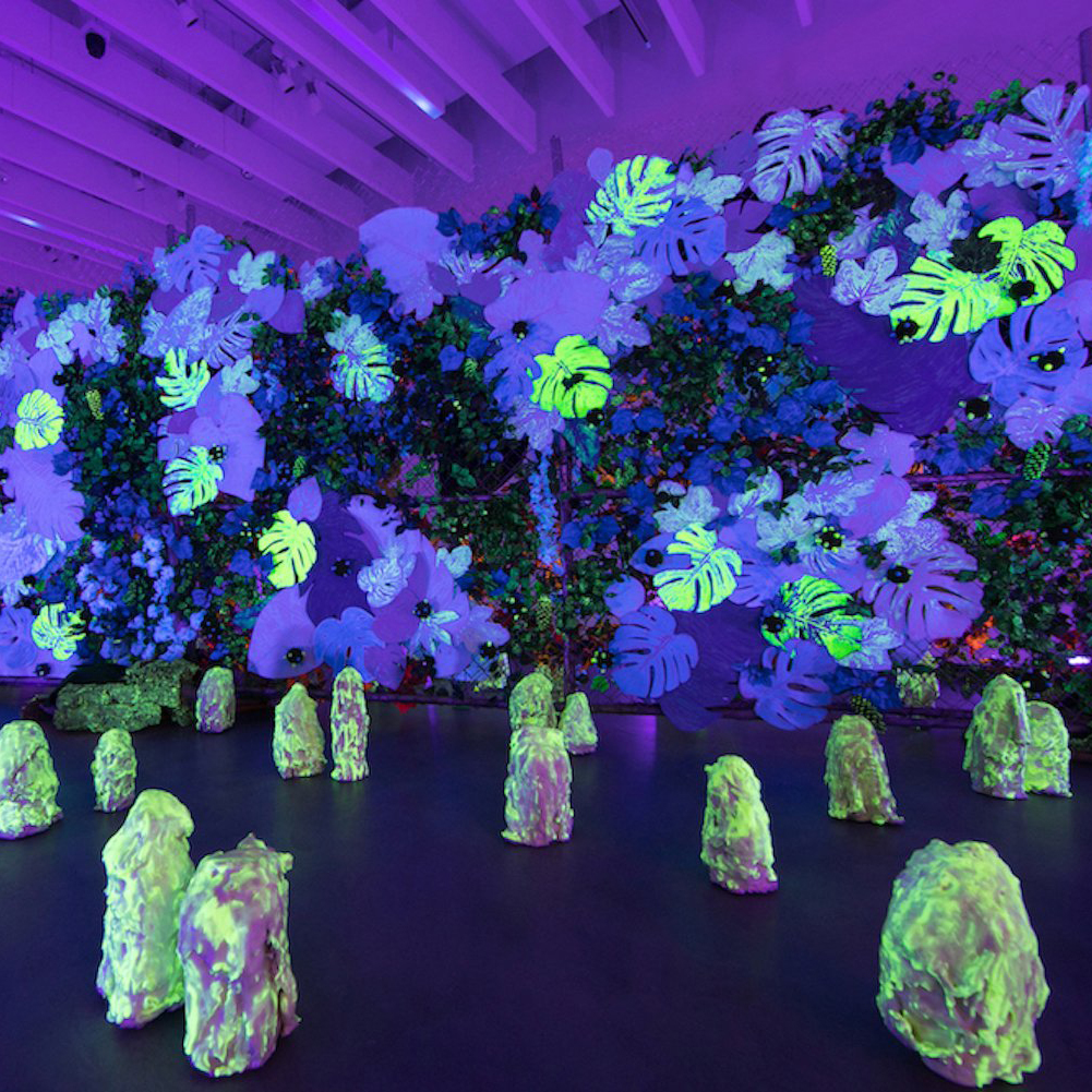 Large immersive installation featuring a wall of layered artificial foliage and flowers glowing under blacklight in shades of neon green, purple, and blue. Bright, fluorescent monstera leaves and blossoms appear to float against a dense backdrop of greenery. On the floor in front, clusters of irregular rock-like forms glow in green and yellow, creating a surreal, otherworldly landscape under the purple-lit ceiling beams of the gallery space.