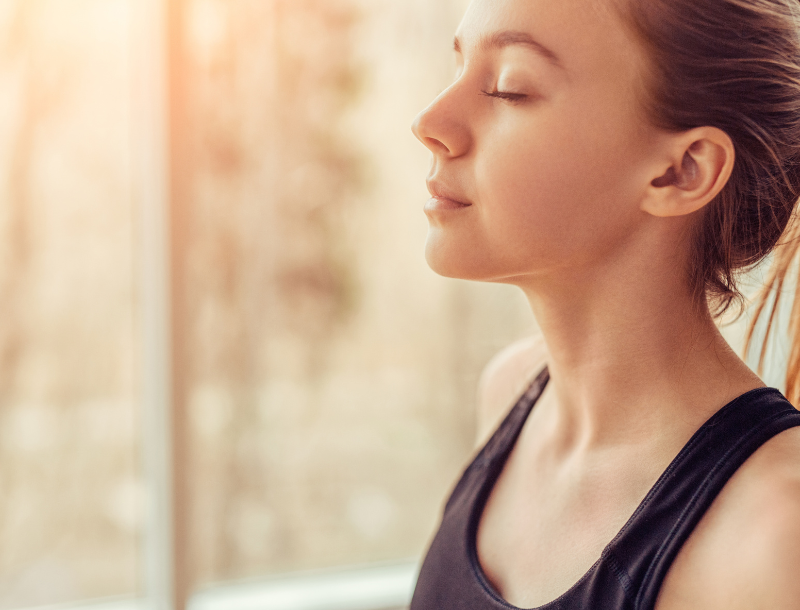 A young woman stands near a window with her eyes closed, softly lit by warm sunlight. She wears a black tank top and appears calm and relaxed, suggesting a moment of meditation or mindfulness.