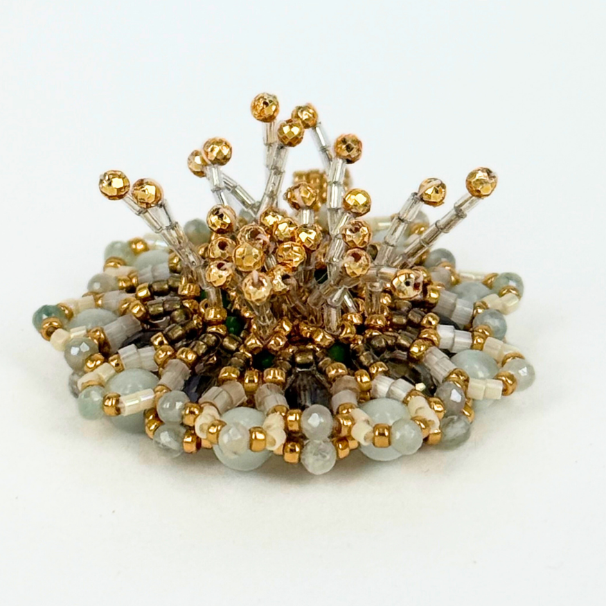 Close-up of a small, sculptural beaded form with pale gray, white, and gold beads arranged in a circular base. Thin bead stems rise upward from the center, tipped with faceted gold beads, creating a delicate, floral or coral-like structure against a white background.
