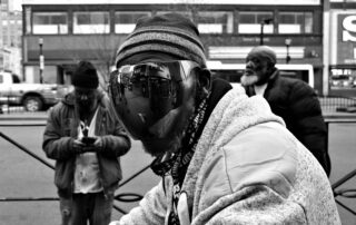 Black-and-white street photograph of three men on a city sidewalk. The man in the foreground wears a striped knit hat and an oversized mirrored visor that reflects storefronts and pedestrians. He leans slightly toward the camera. Behind him, one man looks down at his phone and another stands with hands in pockets, glancing away. Urban buildings and cars are out of focus in the background.