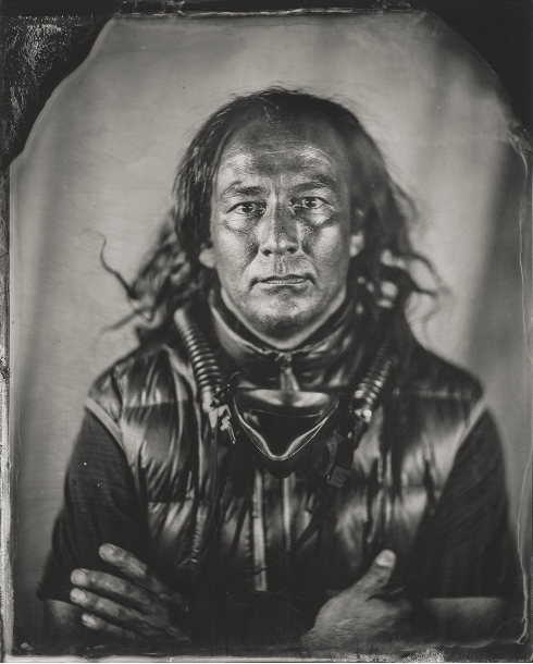 A black-and-white portrait of a man with long hair, looking directly at the camera with a serious expression. He is wearing a shiny, padded vest and has thick cords or breathing tubes draped around his neck and chest. His arms are crossed. The photograph has a wet-plate or tintype style with a soft background and darkened border edges.