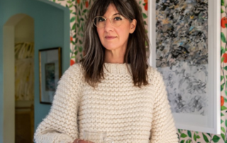 A woman with shoulder-length dark hair and glasses stands indoors holding a clear glass cup. She wears a textured cream-colored sweater and looks slightly off to the side. Behind her are framed abstract artworks on a patterned wall with green leaves and red accents, and an arched doorway leading to another room.