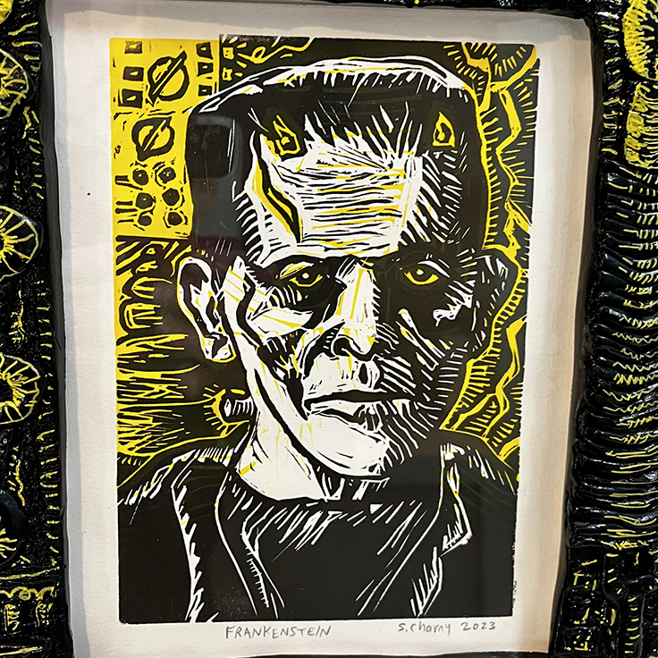 Black-and-yellow linocut-style portrait of Frankenstein’s monster with a stern expression, framed in a thick, textured black frame decorated with raised circular and abstract patterns highlighted in yellow. The print features bold carved lines, yellow eyes, and a graphic yellow background with geometric motifs. The title “FRANKENSTEIN” and the artist’s signature with the year 2023 are visible below the image.