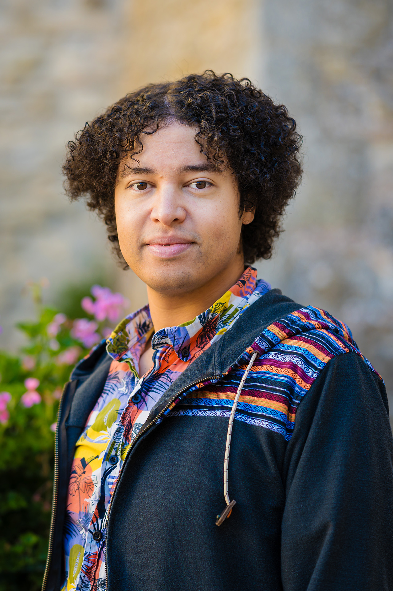 Civitella Ranieri 2025 A young person with dark, curly hair stands outdoors in front of a stone wall, with pink and white flowering bushes visible to the left. They are wearing a black zip-up hoodie over a colorful floral shirt and looking calmly at the camera. The photo is a portrait-style shot taken in warm, natural light.