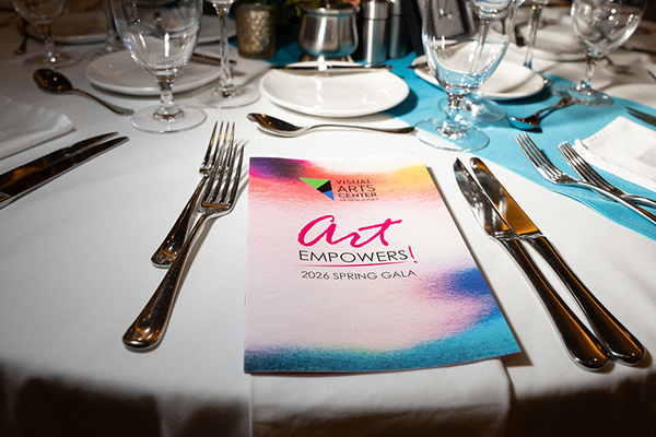 A close-up of an elegantly set round dining table with white linens and teal napkins, featuring a colorful watercolor program booklet reading 'art EMPOWERS! 2026 Spring Gala' from the Visual Arts Center of New Jersey, surrounded by silver cutlery and crystal glassware.